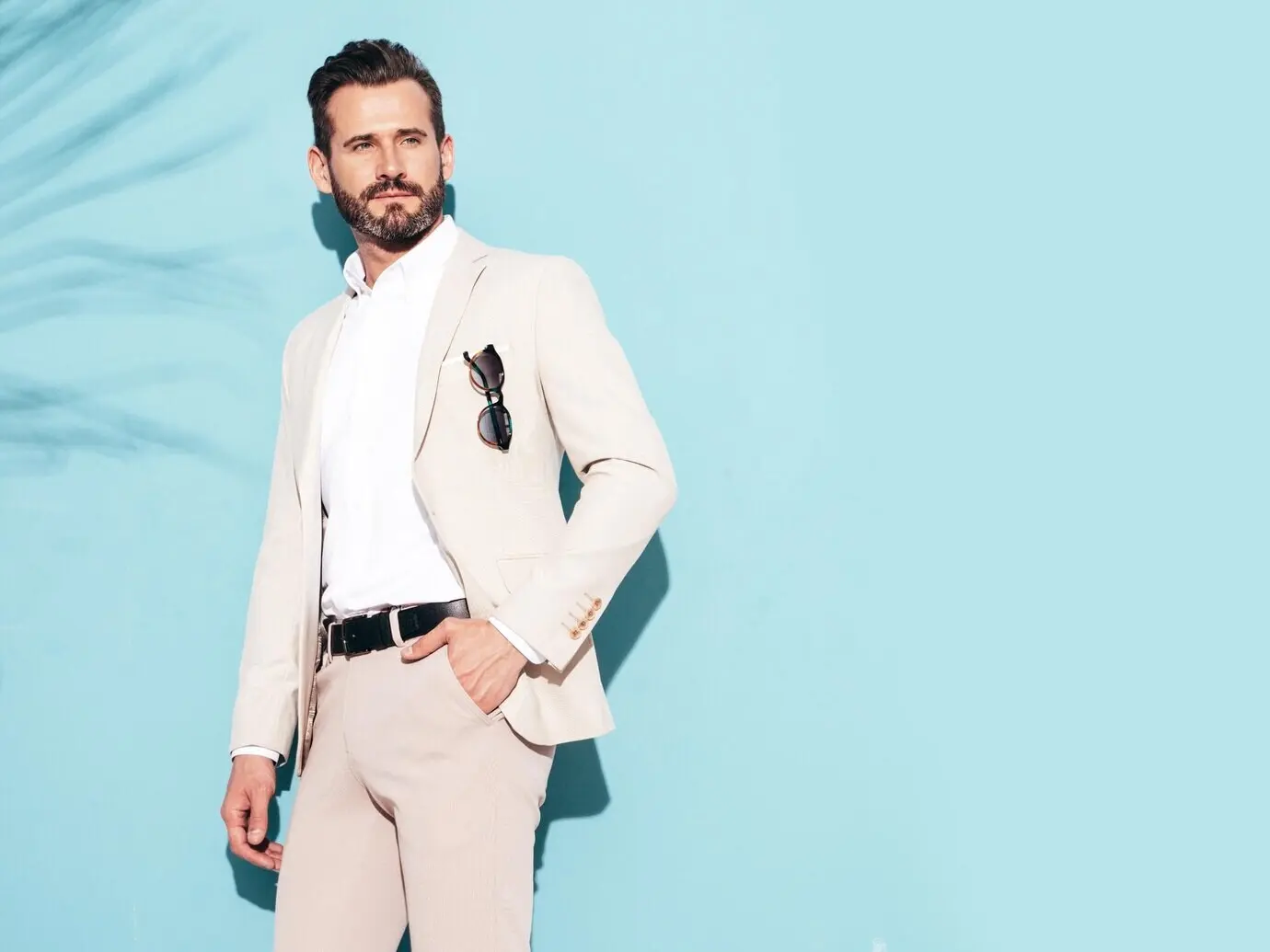 Porträt eines gutaussehenden, selbstbewussten und stilvollen Hipster-Lumbersexual-Models: ein sexy, moderner, modebewusster Mann im eleganten weißen Anzug, der im Studio neben einer blauen Wand mit Sonnenbrille posiert.